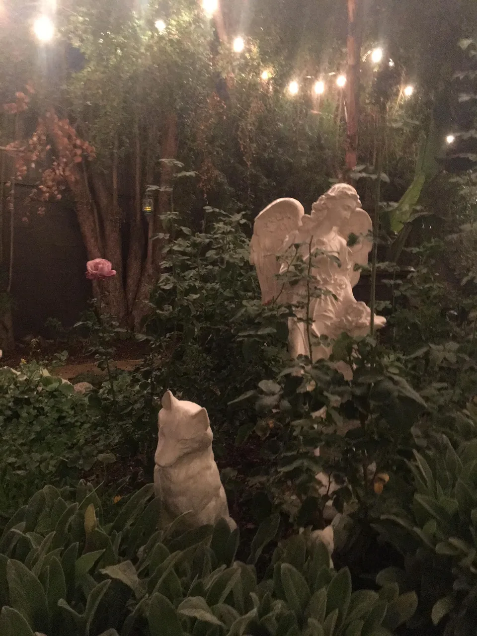 Angel and animal sculptures among plants beneath evening garden lights.